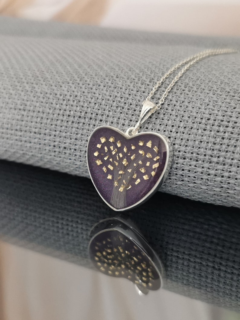 Op de afbeelding: Een zilveren hartvormige ketting met een hanger van paarse hars met een gouden boomontwerp.