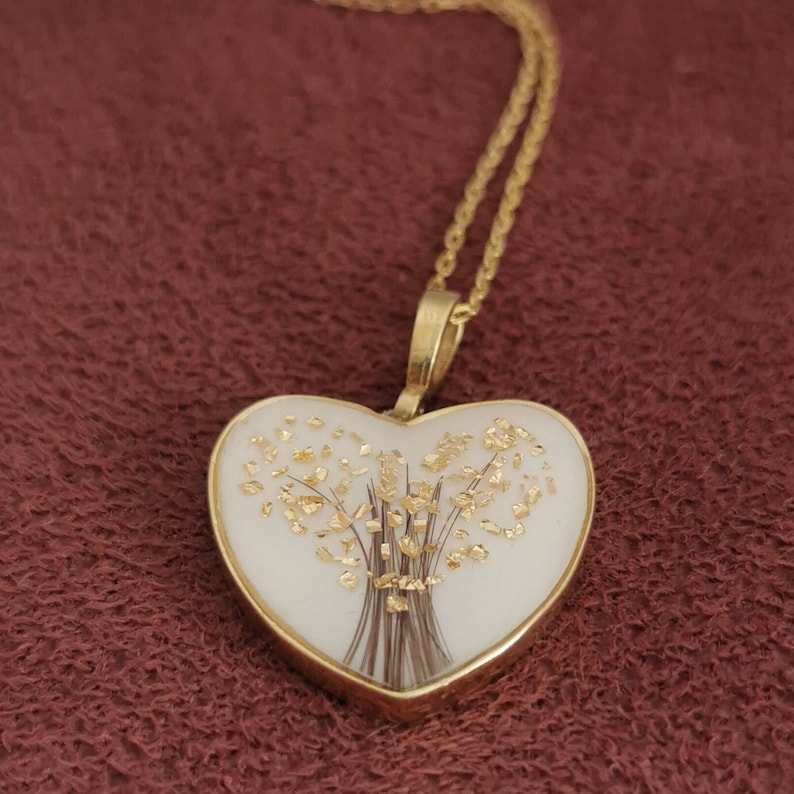 Op de afbeelding: Een gouden hartvormige ketting met een witte hartvormige hanger die een boeket van gouden vlokken en bruin haar bevat.