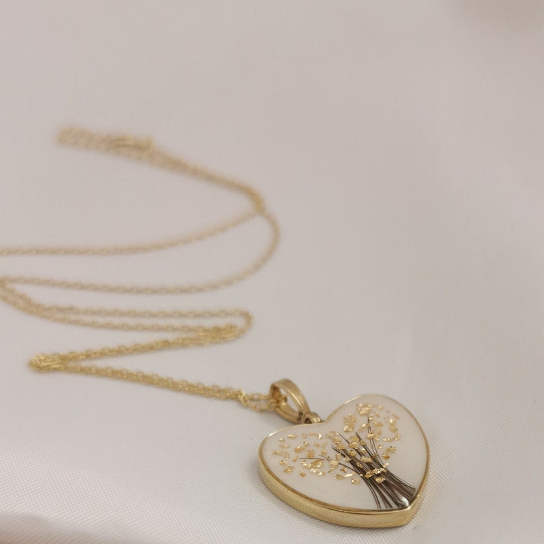 Op de afbeelding: Een gouden ketting met een hartvormige hanger. De hanger is wit met een gouden ontwerp van een boeket bloemen.