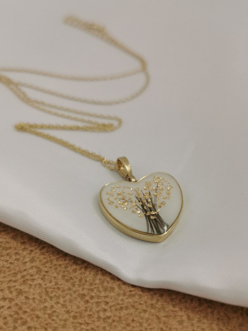 Op de afbeelding: Een gouden ketting met een hartvormige hanger. De hanger is gevuld met helder hars en bevat een gedroogd bloemenboeket met gouden accenten.