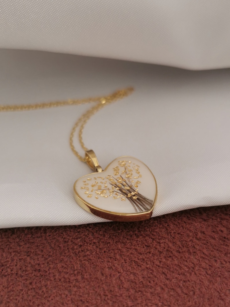Op de afbeelding: Een gouden ketting met een hartvormige hanger, met een witte achtergrond en een delicaat gouden bloemenmotief.