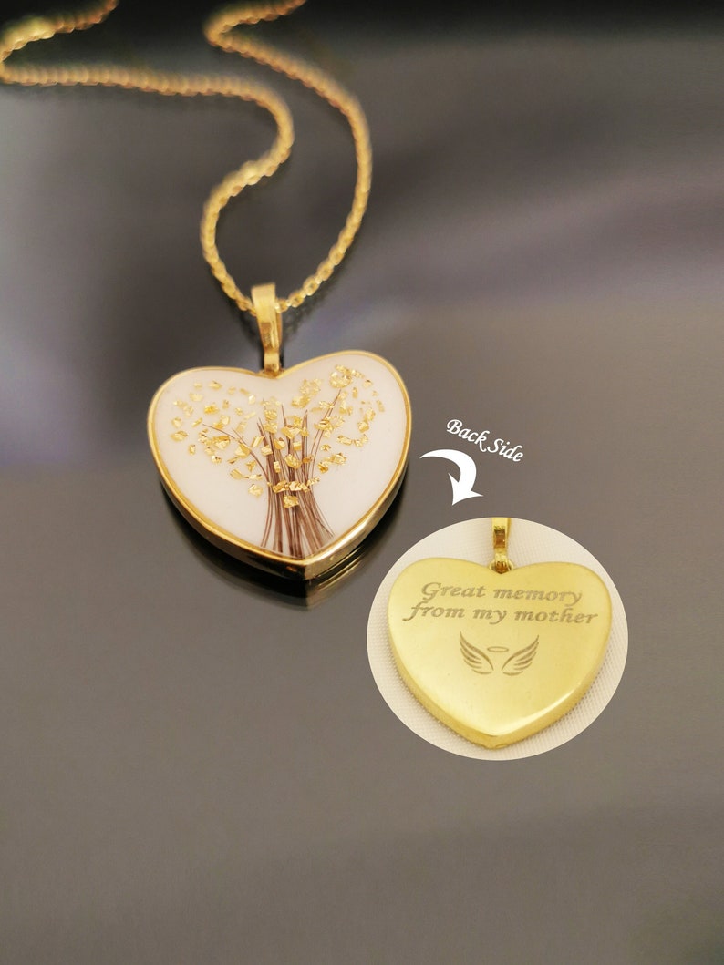 Op de afbeelding: Gouden ketting in de vorm van een hart met een witte hartvormige hanger die gedroogde bloemen en gouden vlokken bevat. De achterkant van de hanger is gegraveerd met de tekst "Great memory from my mother" en een paar engelenvleugels.