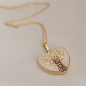 Op de afbeelding: Een gouden ketting met een hartvormige hanger. De hanger is gevuld met witte hars en bevat een gedroogd bloemenboeket met gouden accenten.