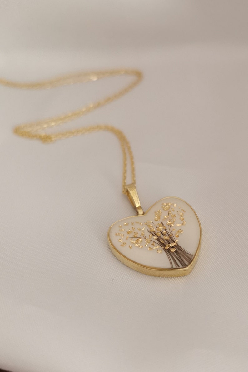 Op de afbeelding: Een gouden ketting met een hartvormige hanger. De hanger is gevuld met witte hars en bevat een gedroogd bloemenboeket met gouden accenten.