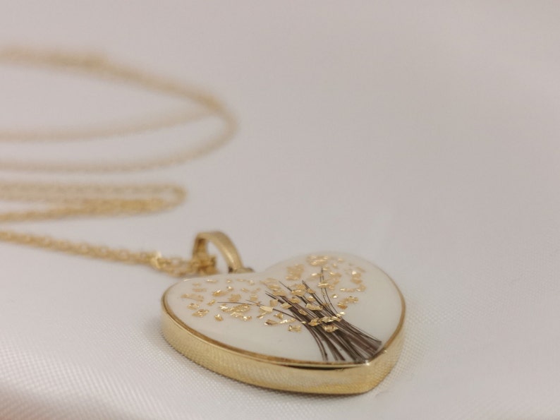 Op de afbeelding: Een gouden hartvormige ketting met een hanger in de vorm van een wit ge&euml;mailleerd hart met een gouden bloemenmotief.