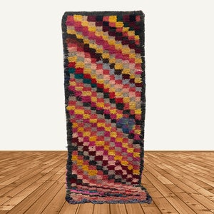 May include: A long, narrow rug with a geometric pattern in shades of pink, yellow, red, blue, and black. The rug has a checkered design with a diagonal pattern.