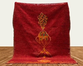 May include: A red Moroccan rug with a traditional geometric pattern in shades of orange, green, and yellow. The rug has a fringed edge and is woven with a high pile.