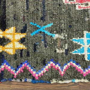 May include: A close-up of a colorful, hand-woven rug with a geometric pattern. The rug is made of various shades of blue, pink, yellow, and white, with a dark gray background.