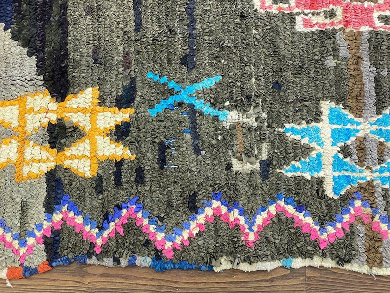 May include: A close-up of a colorful, hand-woven rug with a geometric pattern. The rug is made of various shades of blue, pink, yellow, and white, with a dark gray background.