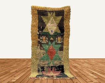 Puede incluir: Una alfombra rectangular hecha a mano con un diseño geométrico central. La alfombra presenta un fondo negro con formas geométricas verdes, rojas y amarillas. El borde es un diseño texturizado y con flecos en tonos amarillos y naranjas. La alfombra se exhibe sobre un suelo de madera.