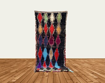 Puede incluir: Una alfombra larga y rectangular con un fondo negro y un patrón de diamantes repetido en varios colores, incluyendo rojo, azul y verde. La alfombra está hecha de un material texturizado y tiene un borde con flecos. La alfombra se muestra sobre un fondo blanco.