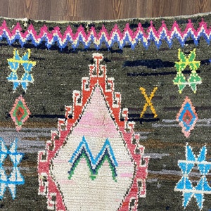 May include: A Moroccan rug with a dark brown background and colorful geometric patterns. The rug features a large diamond shape in the center with a pink, white, and green design. The rug is bordered with a zig-zag pattern in pink, blue, and white.