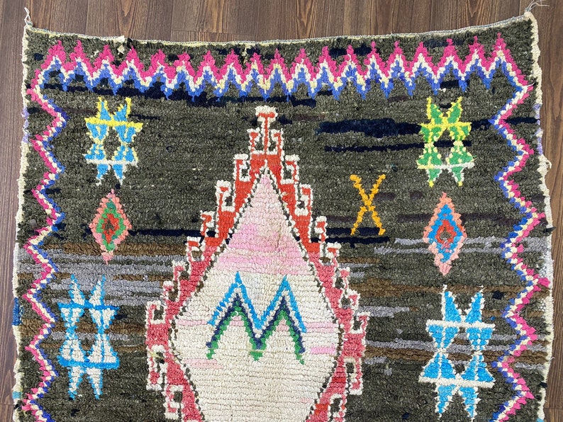 May include: A Moroccan rug with a dark brown background and colorful geometric patterns. The rug features a large diamond shape in the center with a pink, white, and green design. The rug is bordered with a zig-zag pattern in pink, blue, and white.