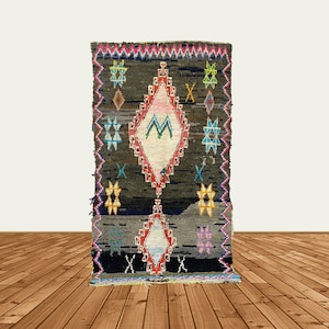 May include: A dark brown rug with a colorful diamond pattern in the center. The diamond pattern features pink, white, blue, and green colors. The rug has a fringe border.