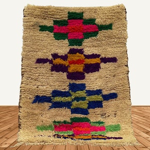 Puede incluir: Una alfombra marroquí beige con un patrón geométrico colorido. El patrón presenta tres filas de cuadrados y rectángulos coloridos en tonos de rosa, verde, azul, amarillo y morado.