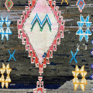 May include: A dark gray rug with a diamond-shaped pattern in the center. The diamond is outlined in red and pink and has a white center with a green and blue design. The rug has a fringe border in pink, blue, and white.