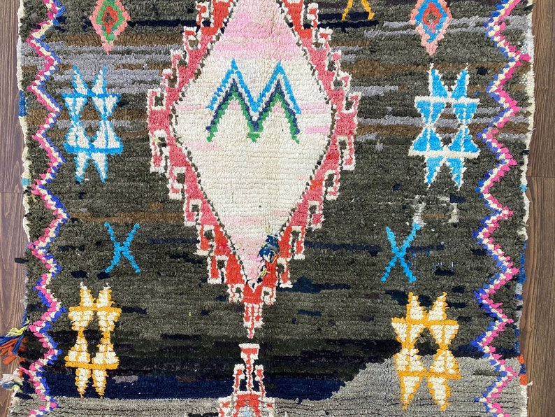 May include: A dark gray rug with a diamond-shaped pattern in the center. The diamond is outlined in red and pink and has a white center with a green and blue design. The rug has a fringe border in pink, blue, and white.