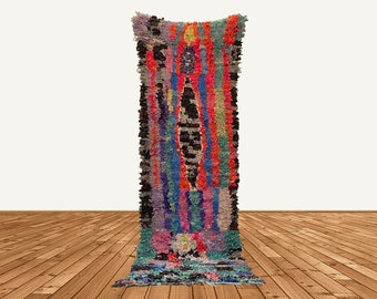 May include: A long, colorful Moroccan rug with a geometric pattern in shades of pink, blue, green, orange, and black. The rug is made of wool and has a hand-woven texture.
