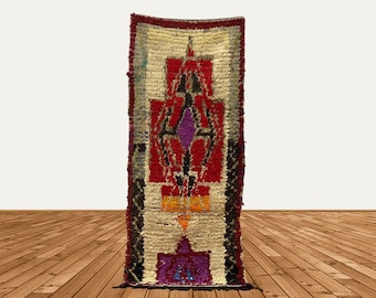 Puede incluir: Una alfombra rectangular larga con un patrón geométrico en tonos de rojo, blanco, negro y morado. La alfombra tiene un borde con flecos y está hecha de un material suave y texturizado.