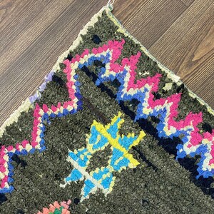 May include: Close-up of a colorful Moroccan rug with a geometric pattern. The rug is made of wool and has a dark green background with pink, blue, yellow, and white accents.