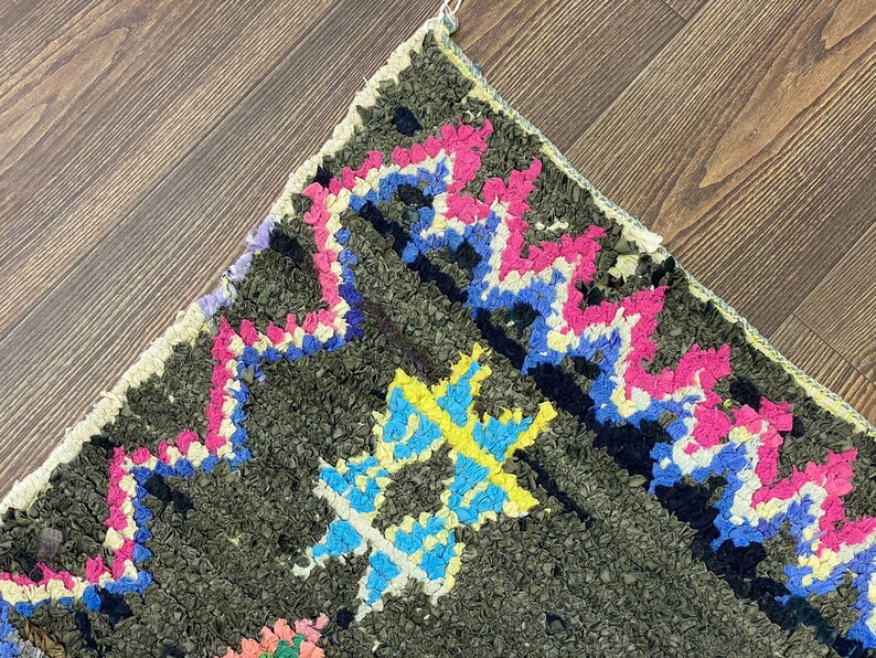May include: Close-up of a colorful Moroccan rug with a geometric pattern. The rug is made of wool and has a dark green background with pink, blue, yellow, and white accents.