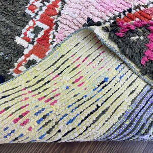 May include: Close-up of a colorful, hand-woven rug with a textured, knotted surface. The rug features a variety of colors, including pink, red, blue, and green. The rug is folded in half, revealing the underside, which is a lighter color with a more simple weave.