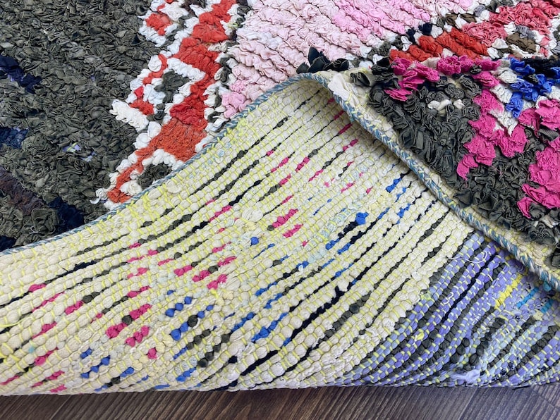 May include: Close-up of a colorful, hand-woven rug with a textured, knotted surface. The rug features a variety of colors, including pink, red, blue, and green. The rug is folded in half, revealing the underside, which is a lighter color with a more simple weave.