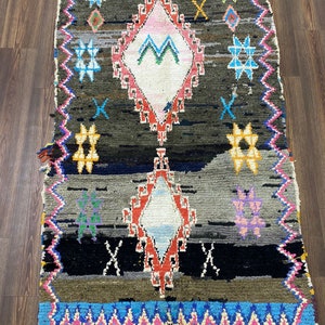 May include: A colorful Moroccan rug with a geometric pattern in shades of red, blue, green, yellow, and pink. The rug has a diamond shape in the center and is surrounded by a border of zigzags and stars.