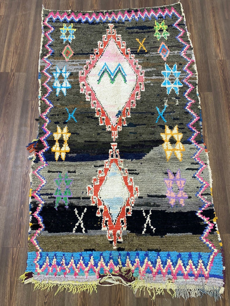 May include: A colorful Moroccan rug with a geometric pattern in shades of red, blue, green, yellow, and pink. The rug has a diamond shape in the center and is surrounded by a border of zigzags and stars.