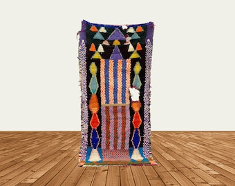 May include: A colorful Moroccan rug with geometric patterns in shades of blue, purple, orange, and red. The rug has a fringed edge and is hanging on a white wall.