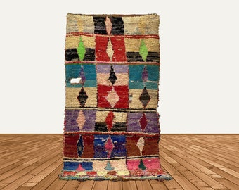 Puede incluir: Una alfombra marroquí colorida con un patrón geométrico de diamantes en tonos de rojo, azul, verde y negro. La alfombra está tejida con una variedad de texturas y materiales, creando un diseño único y llamativo.