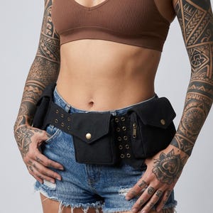 Puede incluir: Riñonera de lona negra con múltiples bolsillos y hebilla de latón. El cinturón se lleva sobre unos shorts vaqueros. La riñonera tiene un bolsillo con cremallera y varios cierres a presión. Accesorio práctico.