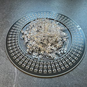 May include: A circular black circuit board with numerous small holes and text, surrounded by a white border. A pile of clear LED lights with metal leads rests in the center. The board is on a dark gray surface.