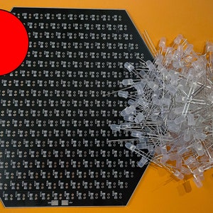 May include: A black hexagonal circuit board with a red circle in the upper left corner. The board is covered in small, round holes. A pile of clear LED lights with silver leads is in the lower right corner.