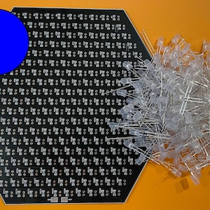May include: A black hexagonal circuit board with a blue circle in the upper left corner. A large pile of clear LED lights are to the right of the board.