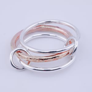 May include: A set of three interlocking rings. Two rings are silver, and one is rose gold. The rose gold ring has a hammered texture. The rings are intertwined, creating a unique and stylish design. The rings are likely jewelry.
