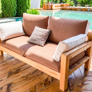 May include: A wooden outdoor loveseat with brown cushions and white pillows. A patterned throw pillow adds a decorative touch. The loveseat is placed on a wooden deck near a swimming pool, creating a relaxing outdoor seating area.