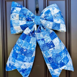 Puede incluir: Un gran lazo decorativo construido a partir de un mosaico de varios cuadrados de tela con patrones florales en azul y blanco. El lazo presenta dos bucles superiores anchos y dos colas largas y puntiagudas, todo acolchado con un patrón de rejilla.