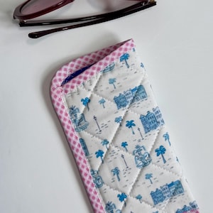 May include: A pair of burgundy sunglasses rests above a quilted eyeglass case. The case features a white background with a blue toile pattern of buildings and palm trees, framed by a pink gingham border.