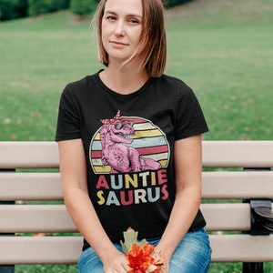 May include: A black t-shirt featuring a pink dinosaur wearing sunglasses and a red bandana. The dinosaur is set against a retro-style sunset with yellow, orange, and blue stripes. The text "Auntie Saurus" is printed below the dinosaur.