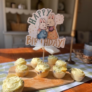 Może przedstawiać: Drewniana deska zawiera babeczki z żółtym lukrem. Ozdobny znak z napisem "Happy Birthday!" i ilustracjami dwóch królików umieszczony jest na środku. Babeczki ułożone są na zielono-białym, w kratę obrusie.