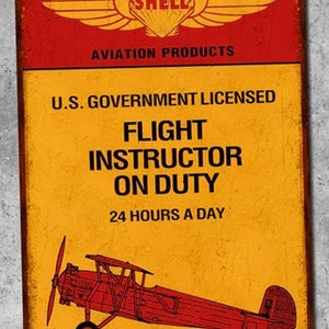 Peut inclure: Une plaque métallique vintage jaune et rouge avec le logo Shell et le texte "Aviation Products". La plaque indique également "U.S. Government Licensed Flight Instructor On Duty 24 Hours A Day" et présente un avion Boeing F3B-1 rouge et blanc.