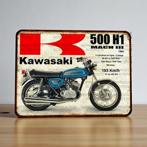 Op de afbeelding: Vintage Kawasaki 500 H1 Mach III motorbord. Het bord toont een blauwe motorfiets met zwarte accenten, het Kawasaki-logo en tekst. Het bord heeft een verweerde, verouderde uitstraling.