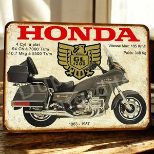 May include: A vintage-style metal sign featuring a motorcycle illustration. The sign has a distressed, aged appearance with the word "HONDA" in red at the top. Details include "GL 1200" and specifications like "185 Km/h" and "348 Kg". The years "1983-1987" are also displayed.