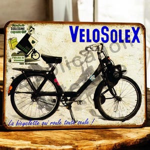 May include: Aged metal sign depicting a black VeloSolex bicycle. The sign features the words "VELOSOLEX" in blue, with additional text including "Moubelle SOLEXINE capsule Or" and "La bicyclette qui roule toute seale!" The sign has a distressed finish.