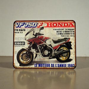 Könnte beinhalten: Ein Vintage-Metallschild mit einer rot-weißen Honda VF750F Motorrad. Das Schild enthält französischen Text, wie z. B. „LE MOTEUR DE L'ANNEE 1983“ und Spezifikationen wie 216 Km/h und 400 DA: 12,3. Das Schild hat ein gealtertes Aussehen.