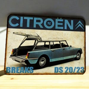 May include: A distressed metal sign depicting a blue Citroën DS 20/23 estate car with an open tailgate. The sign features the word "CITROEN" in blue, along with "BREAKS" and the year "72". The overall design has a vintage aesthetic.