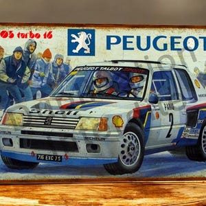 Peut inclure: Panneau métallique vintage représentant une Peugeot 205 Turbo 16 blanche avec des détails bleus et rouges. Le panneau indique "205 turbo 16" et "PEUGEOT". La voiture porte le numéro 2 sur le côté.