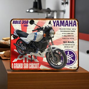 Peut inclure: Panneau métallique rectangulaire avec une moto Yamaha RDLC 350 vintage. Le fond est rouge et blanc avec un motif de drapeau japonais. La moto est blanche et noire avec des détails bleus. Le texte comprend les spécifications et le logo Yamaha.