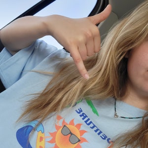 May include: A person wearing a light blue t-shirt with a graphic of a sun wearing sunglasses and riding a surfboard on a wave. The text "303 CULTURE" is printed on the shirt.